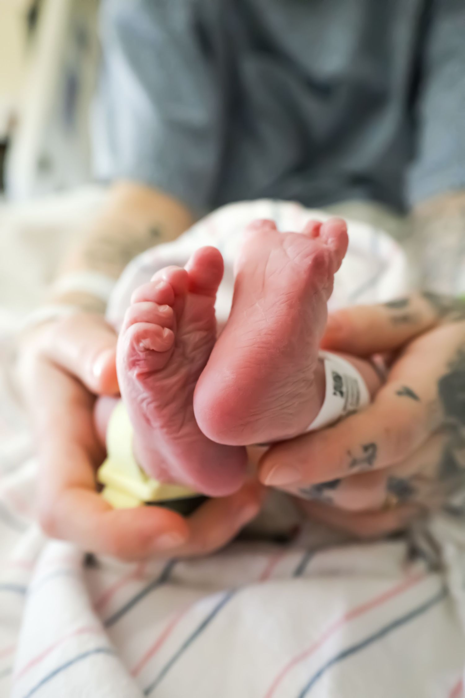 Tattooed hands gently cradle tiny newborn baby feet with hospital ID bracelet after birth.