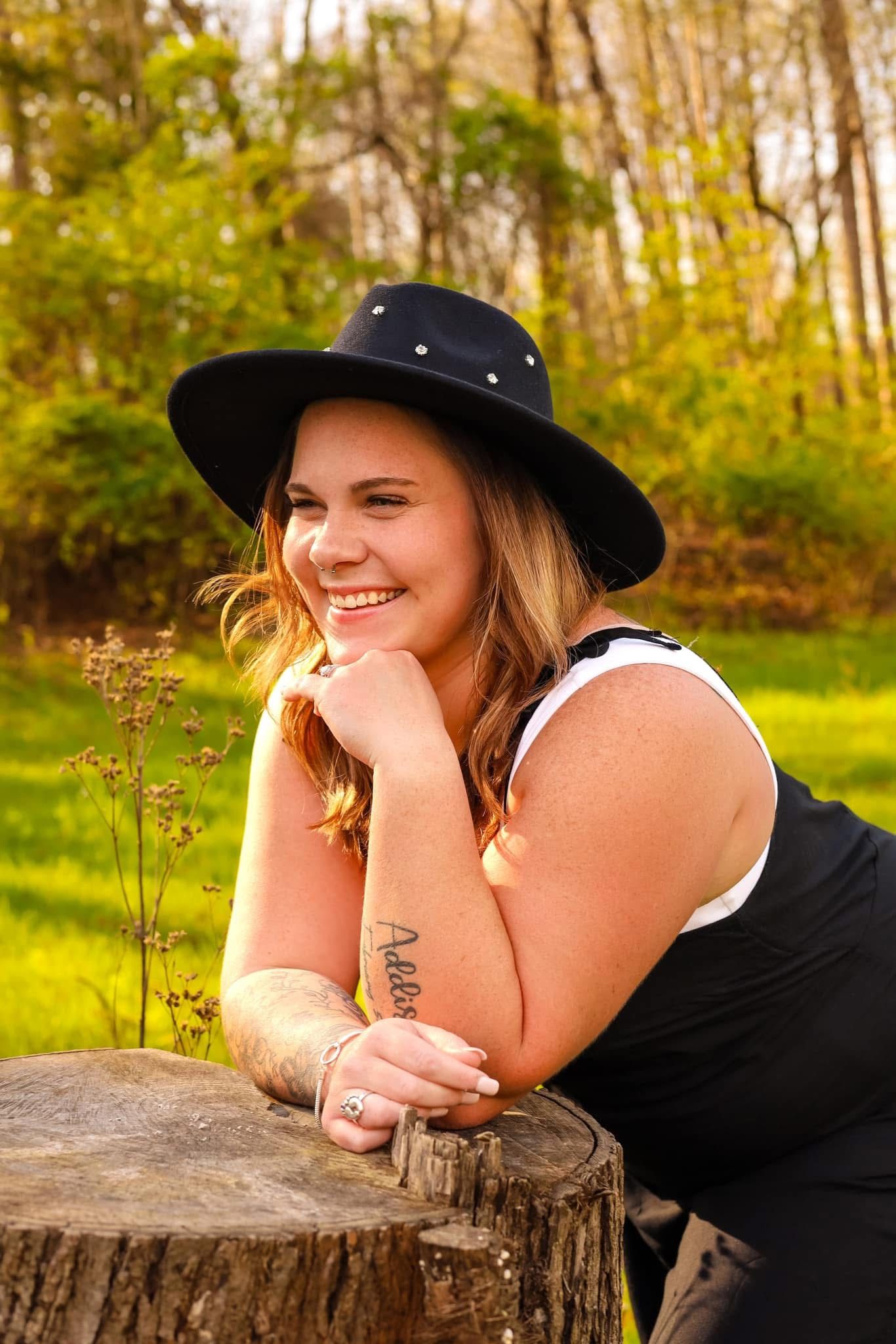 Smiling young woman in black hat and tank top leaning on a tree stump in a sunny green park.