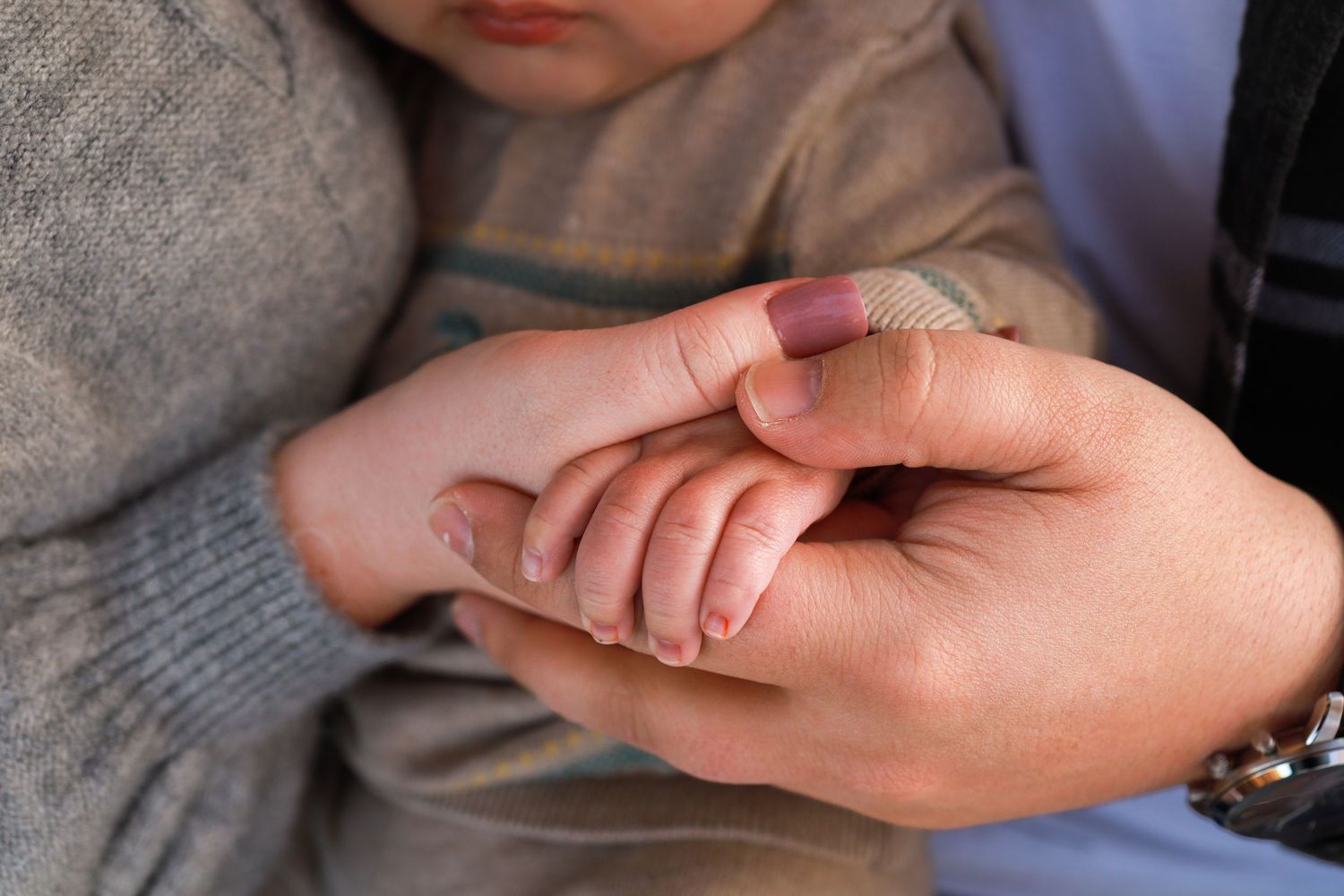 Tiny newborn hand grasping adult finger, baby wearing beige knit sweater in soft natural light.