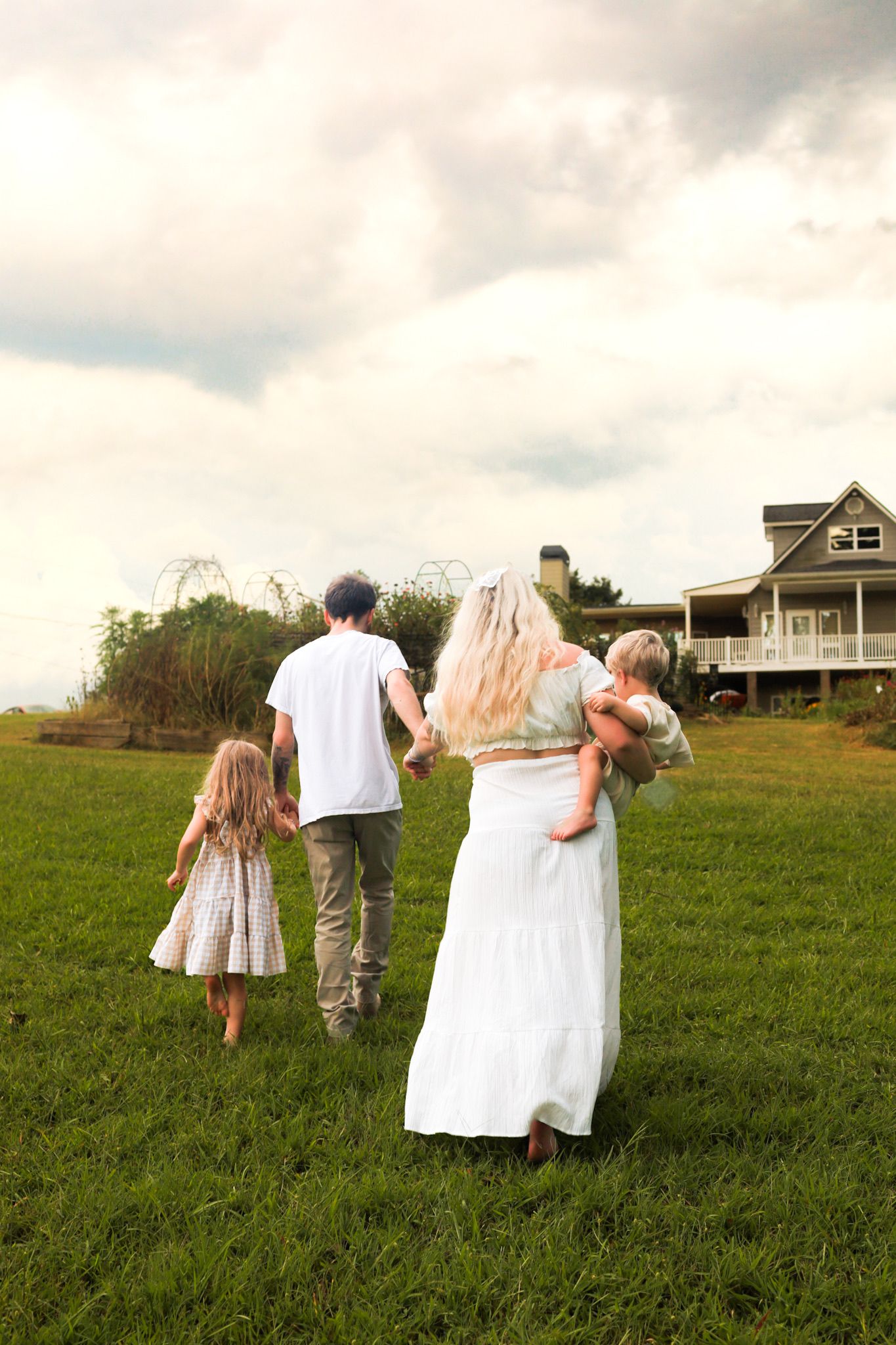 Family of four walking toward large house on green field, mother carrying toddler under cloudy sky.