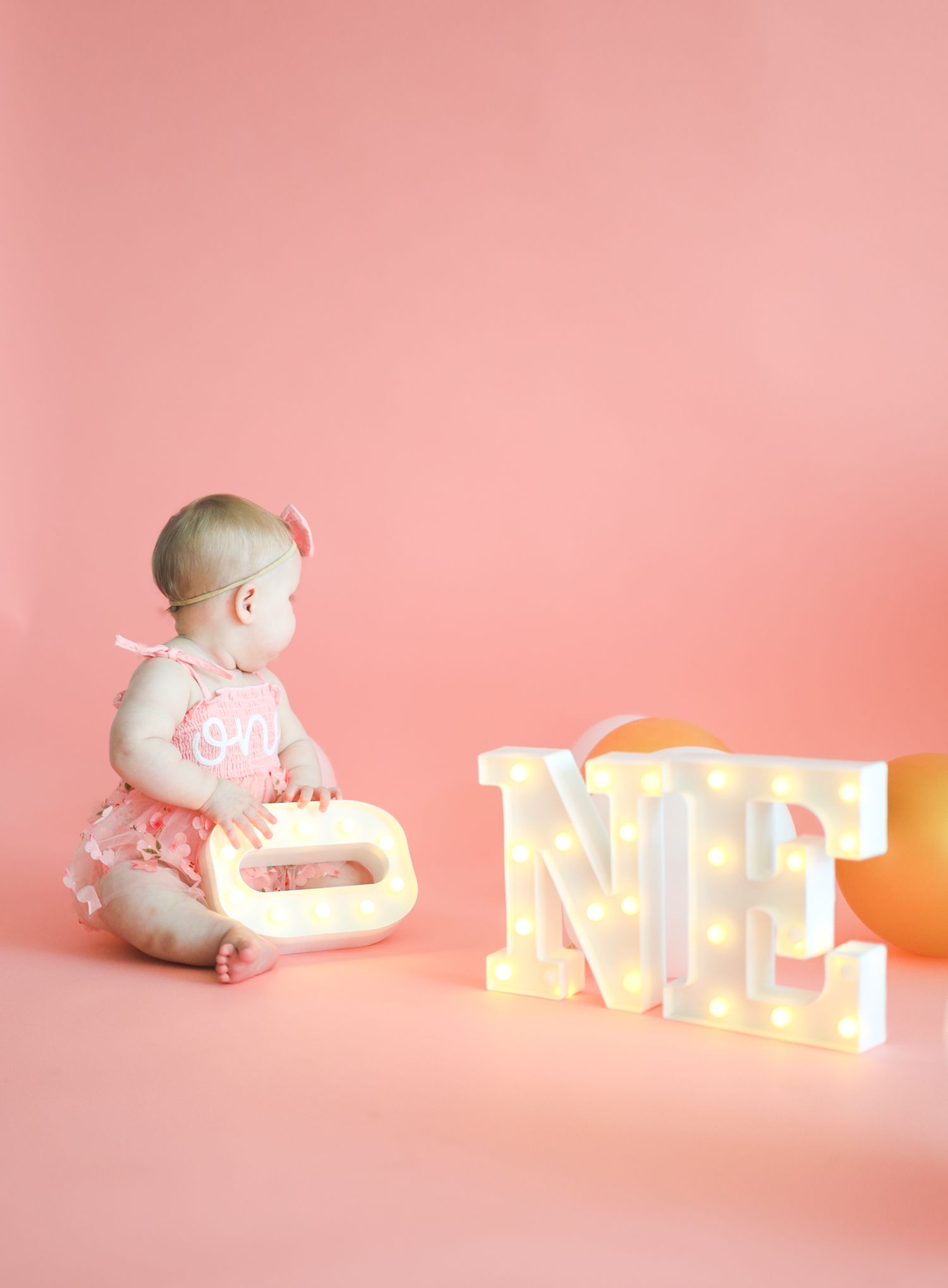 Baby girl in pink tutu sitting beside illuminated ONE marquee letters on pink background.