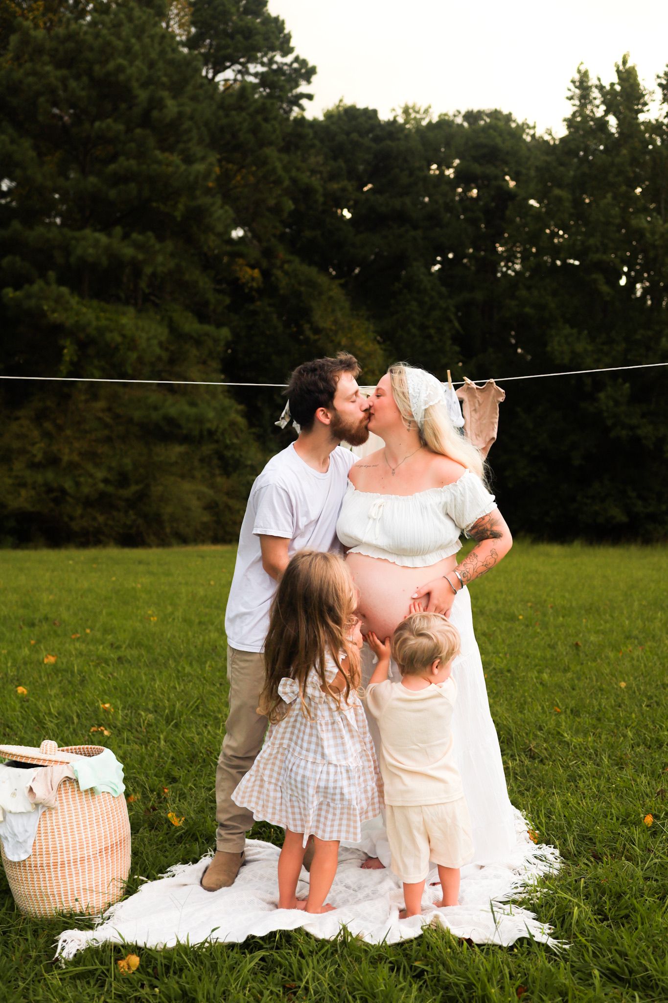Pregnant couple kisses while two young children hug moms belly near clothesline outdoors.