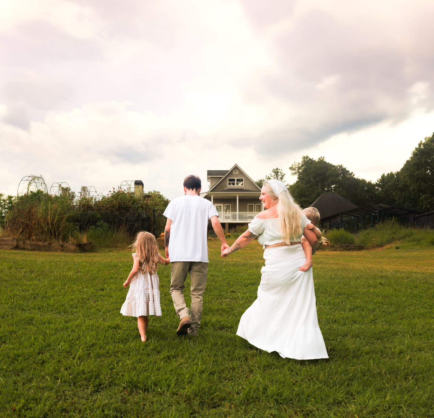 Family of four walking hand-in-hand across green lawn toward large house under cloudy sky.