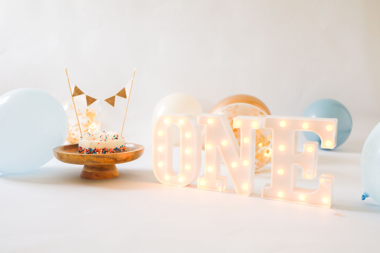 First birthday party setup with smash cake on wooden stand beside illuminated ONE marquee letters and balloons.
