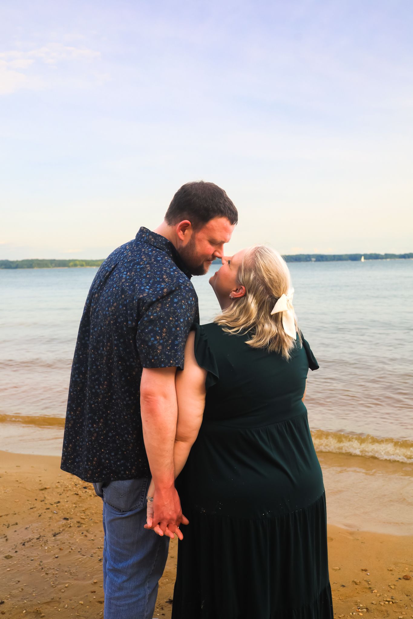 Couple kissing and holding hands on sandy beach with calm blue water in background.