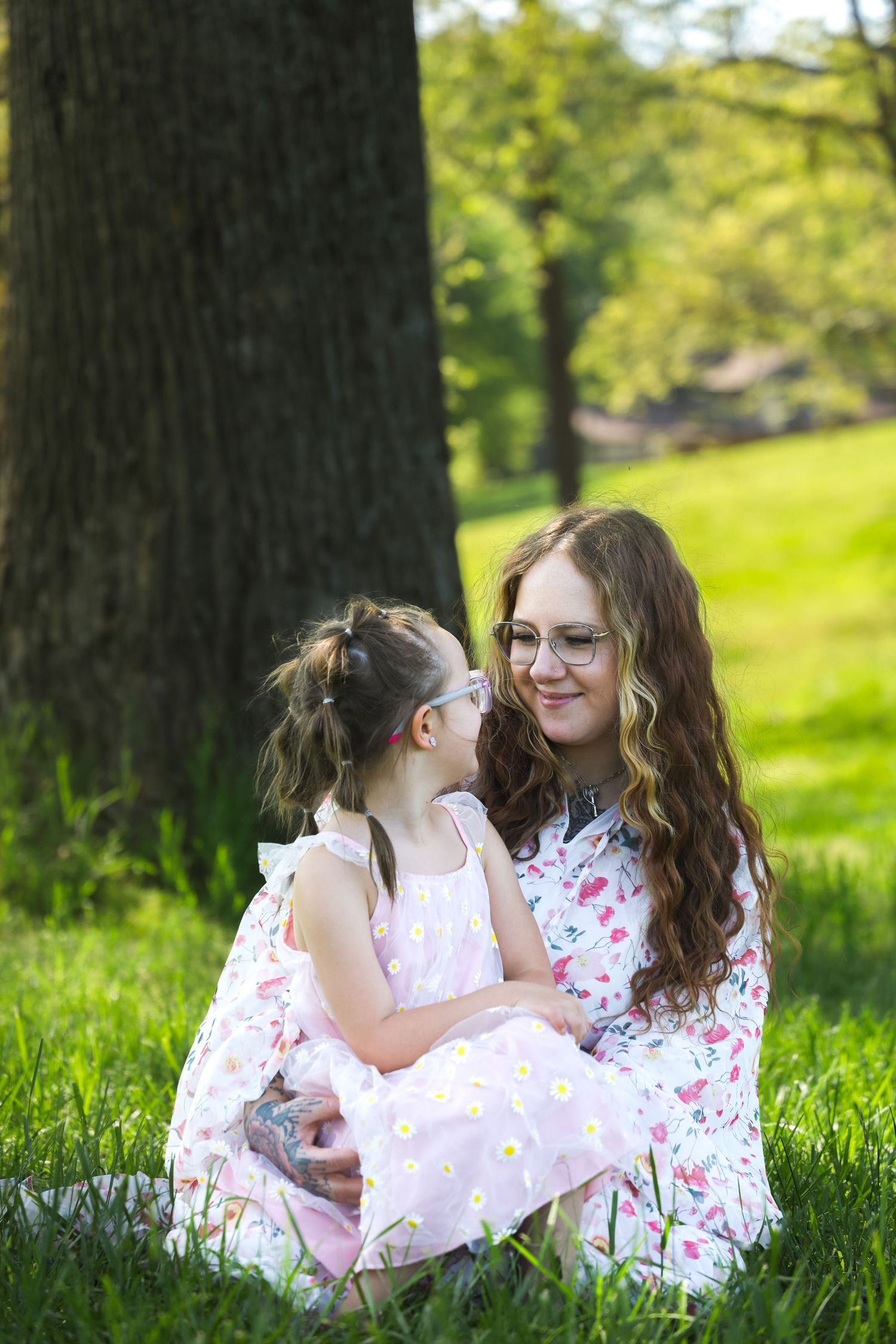 Young girl kissing smiling woman wearing glasses on the cheek while sitting on green grass.