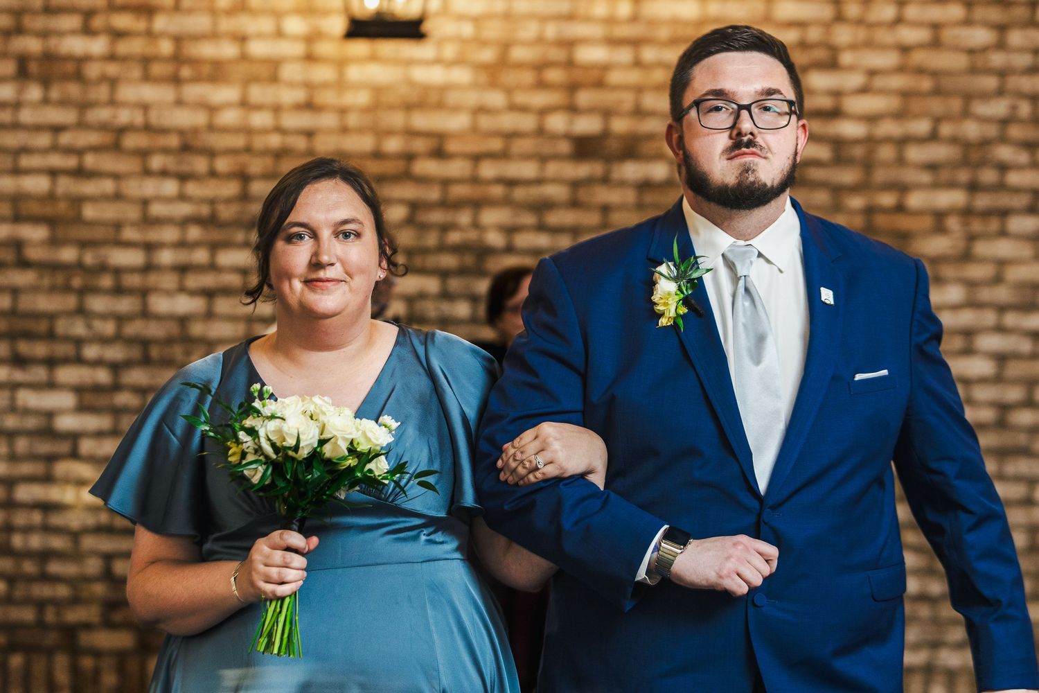Wedding party members in navy blue suits and light blue dresses pose together against a brick wall backdrop.