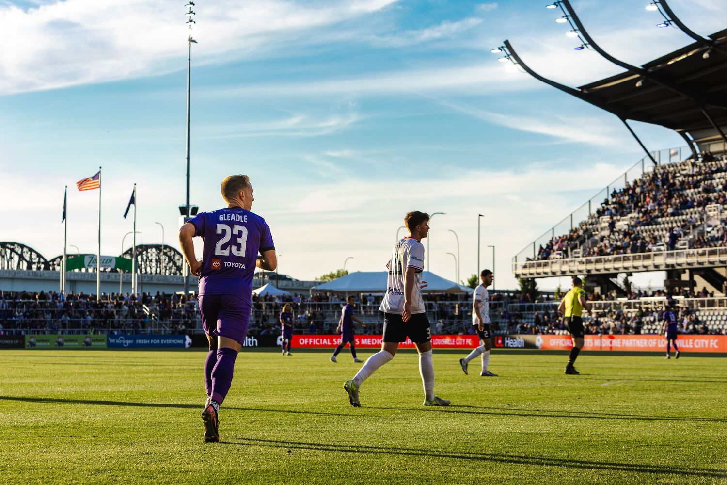 Soccer players on field during match with stadium stands in background.