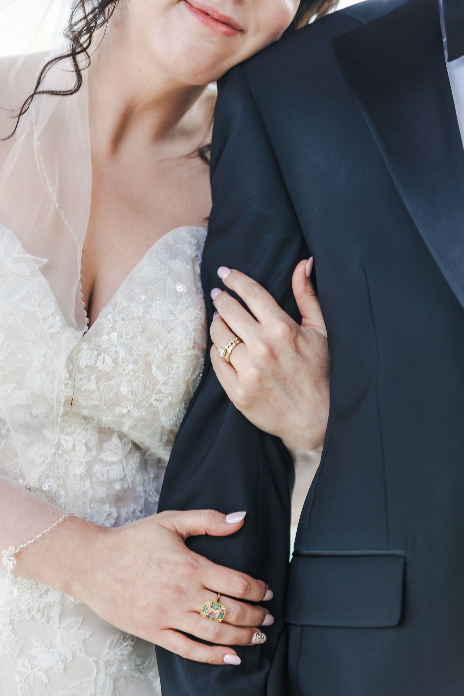 Close-up of bride in lace dress holding groom's arm, showcasing gold diamond engagement and wedding rings.