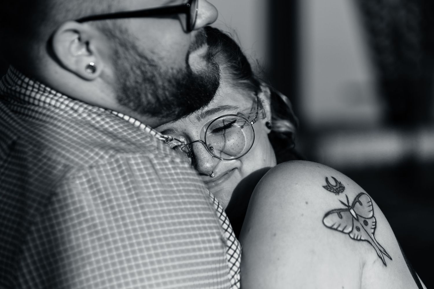 Black and white photo of a couple embracing, woman with butterfly tattoo on shoulder wearing round glasses.