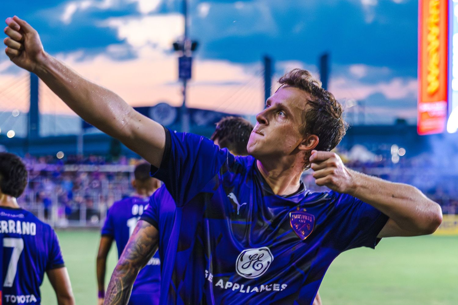 Soccer player in blue jersey celebrates with arms raised during evening match at stadium.