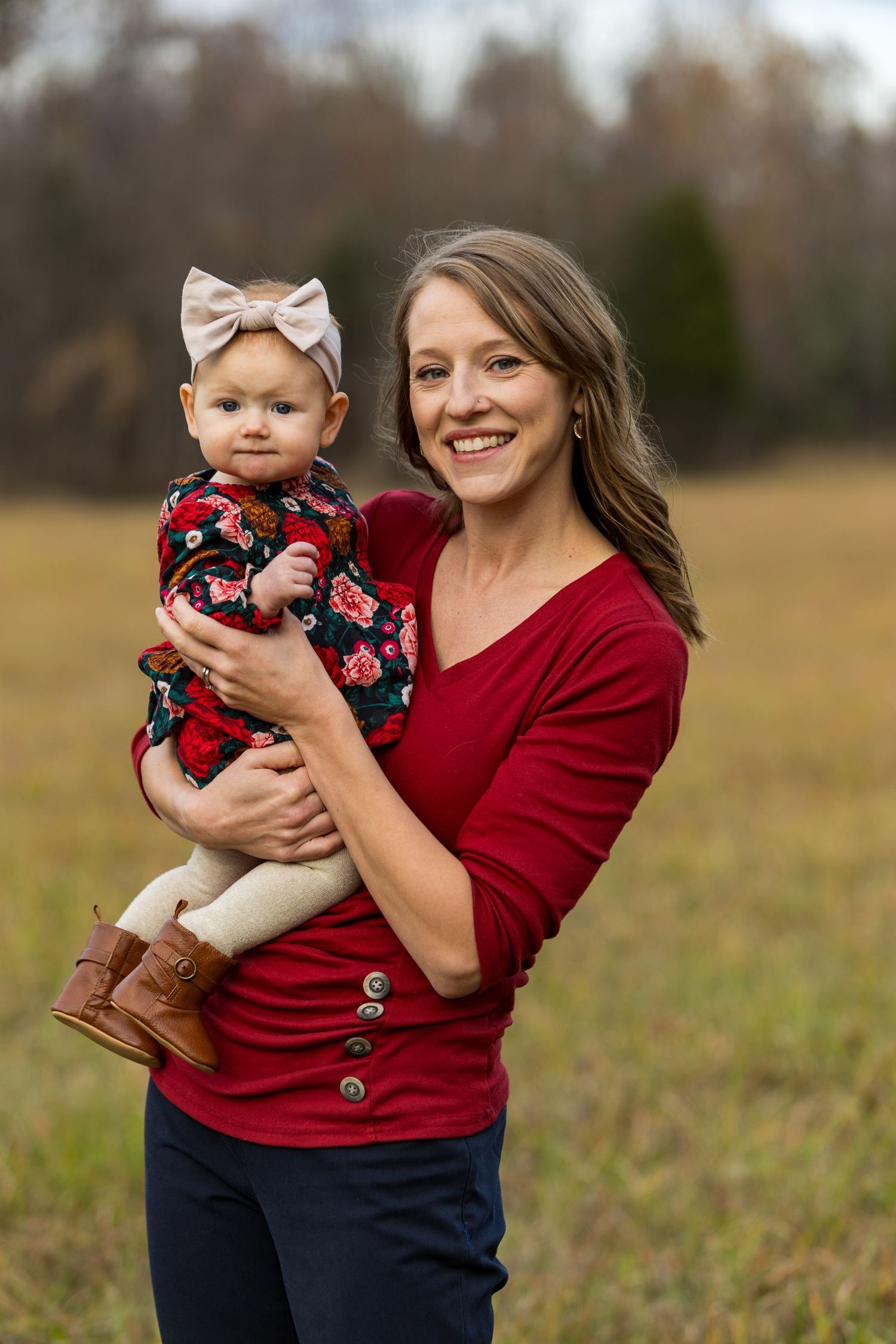 Parent in red sweater holding baby in floral outfit and bow headband outdoors.