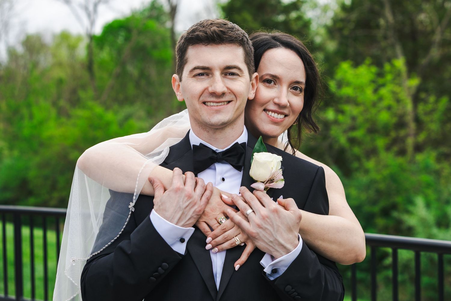 Happy newlywed couple posing outdoors, groom in black tuxedo with boutonniere, bride in white veil, green trees background.