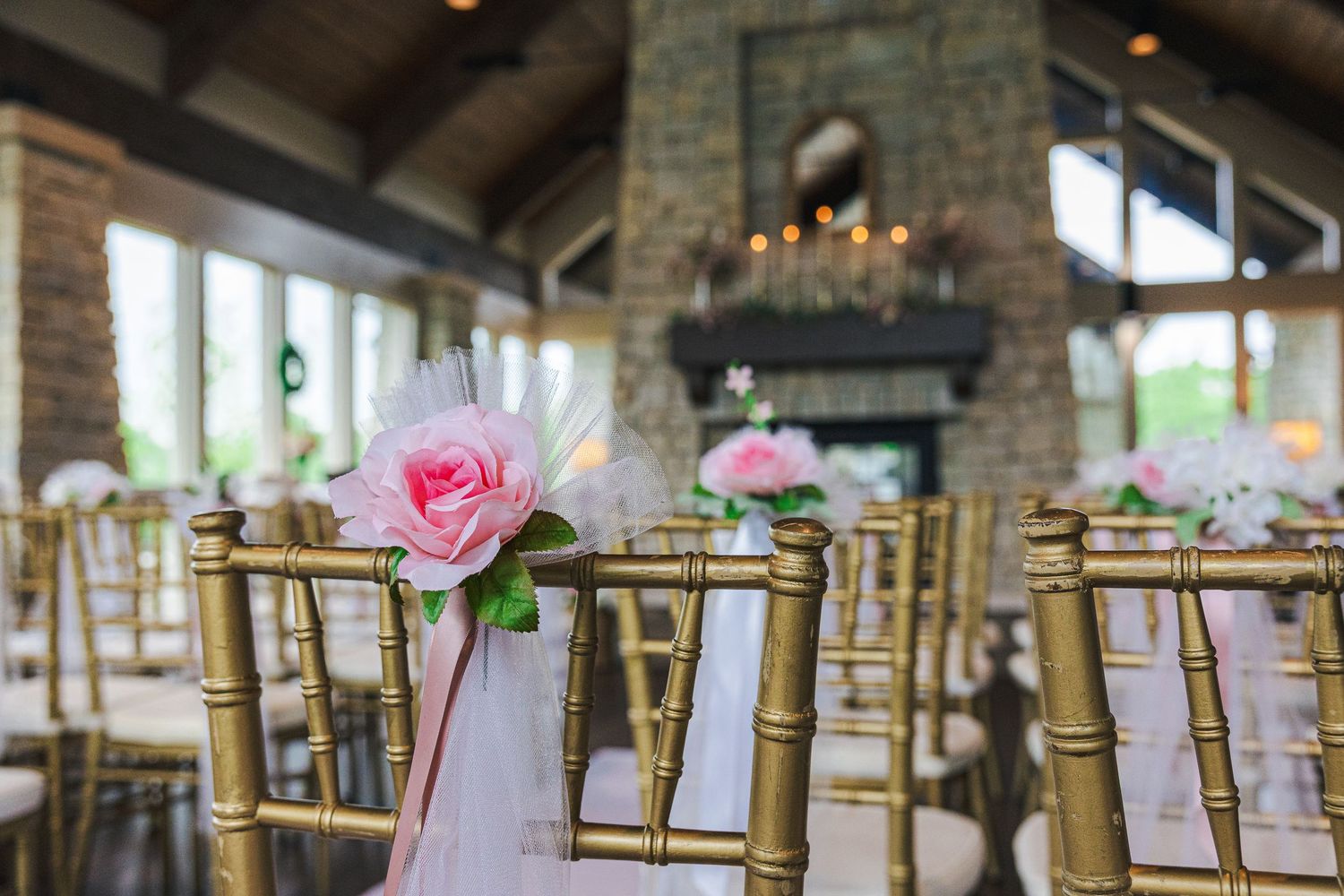Gold chiavari chairs with pink rose and tulle decorations in an elegant wedding venue with stone fireplace.