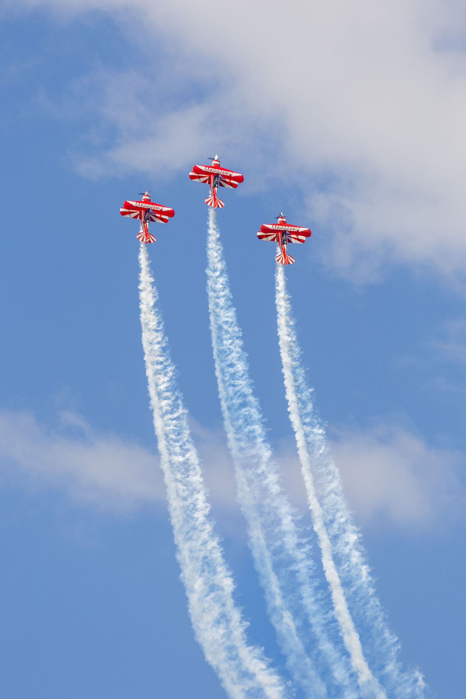 Three red aerobatic planes flying in formation leaving white smoke trails against blue sky.