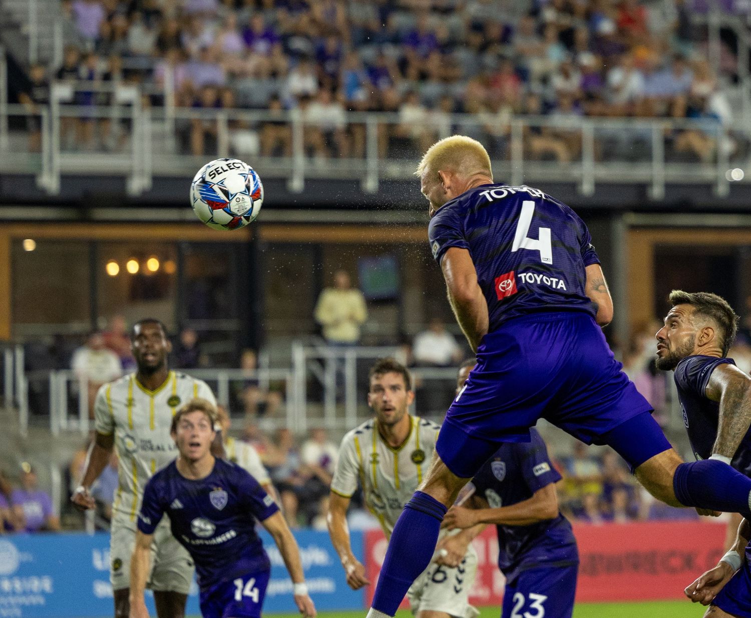 Players compete for header during intense soccer match under stadium lights.