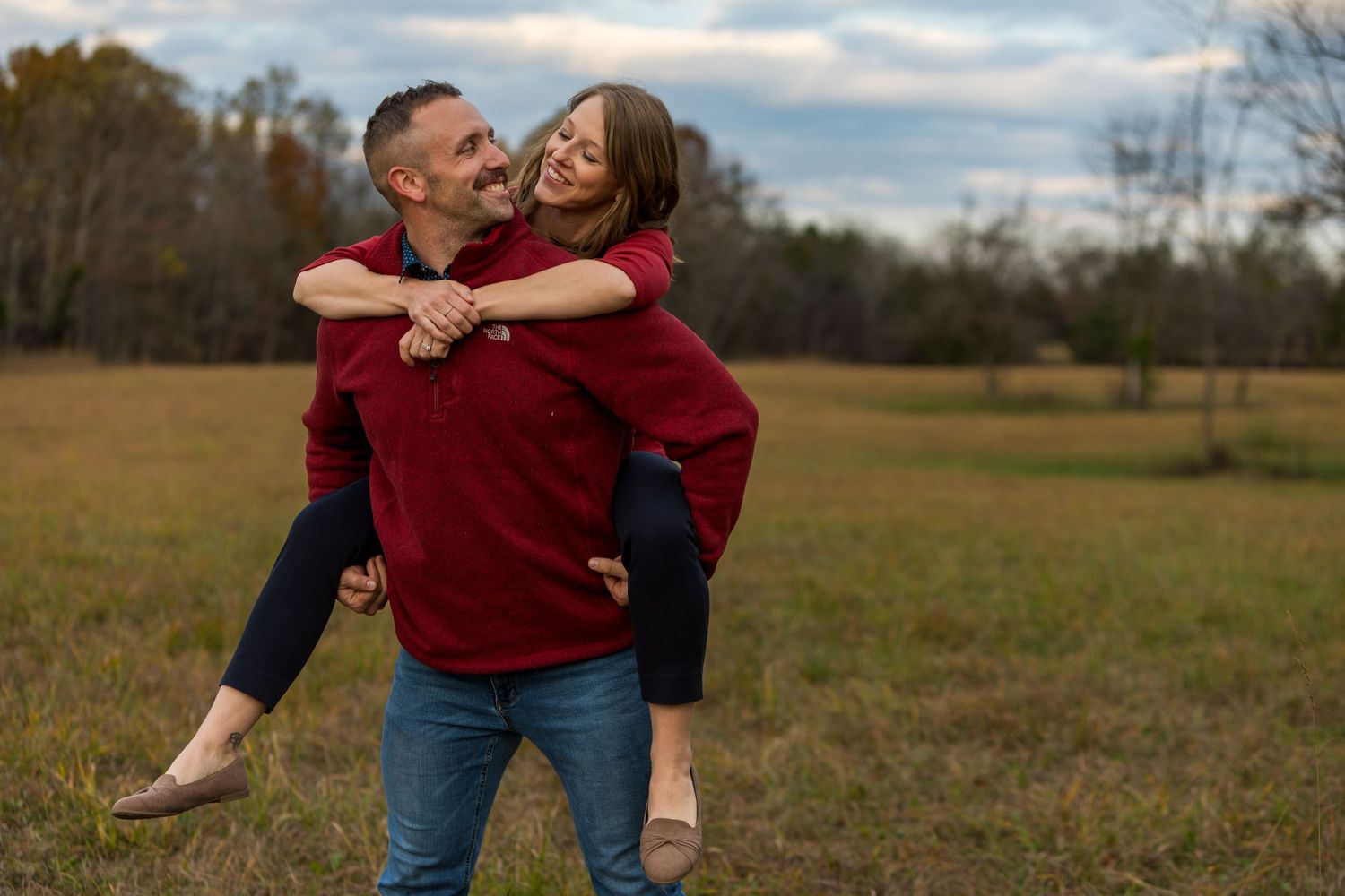 Playful couple in matching red sweaters having fun with piggyback ride in autumn field.