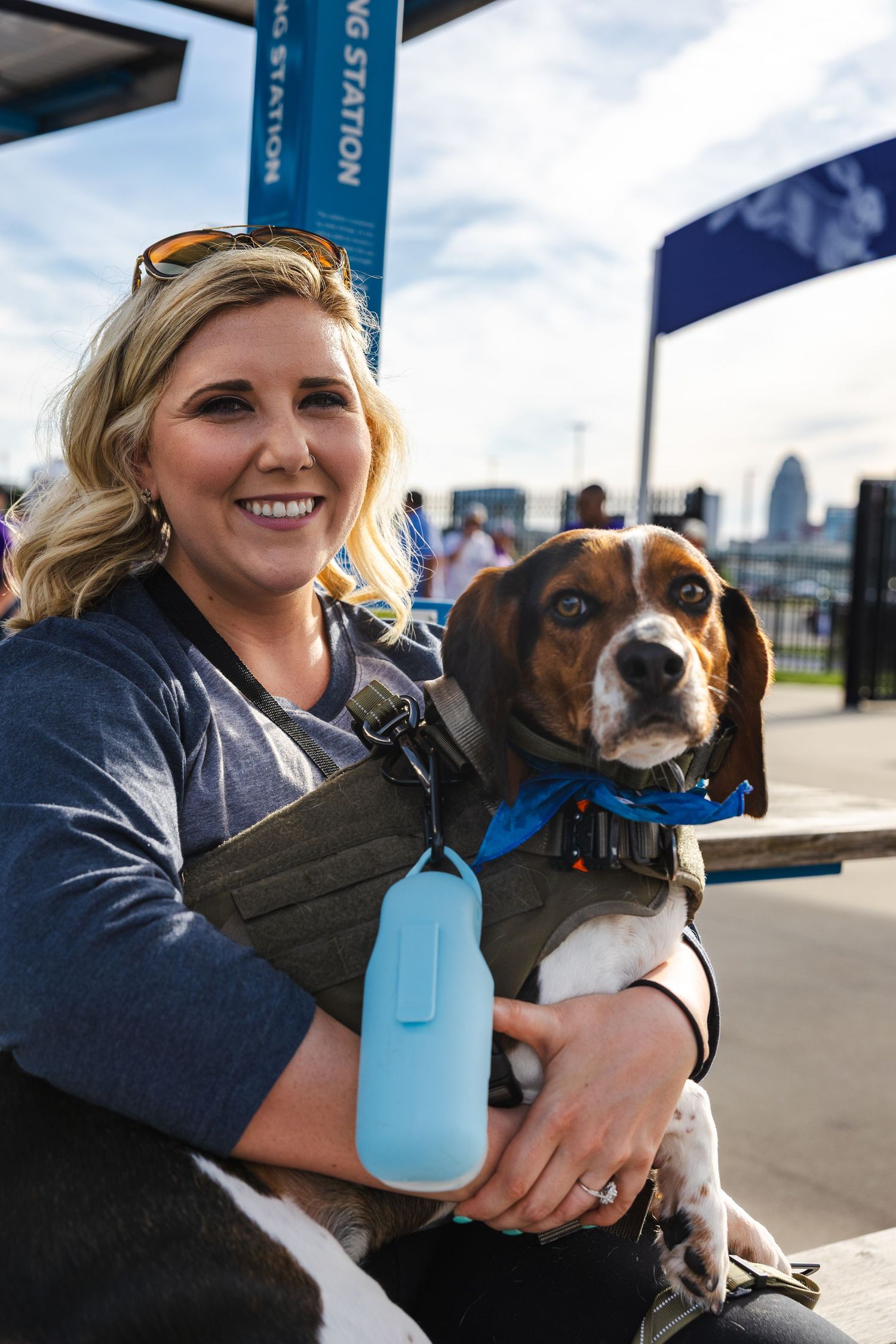 Person holding a beagle mix dog wearing a blue harness outdoors in an urban setting.