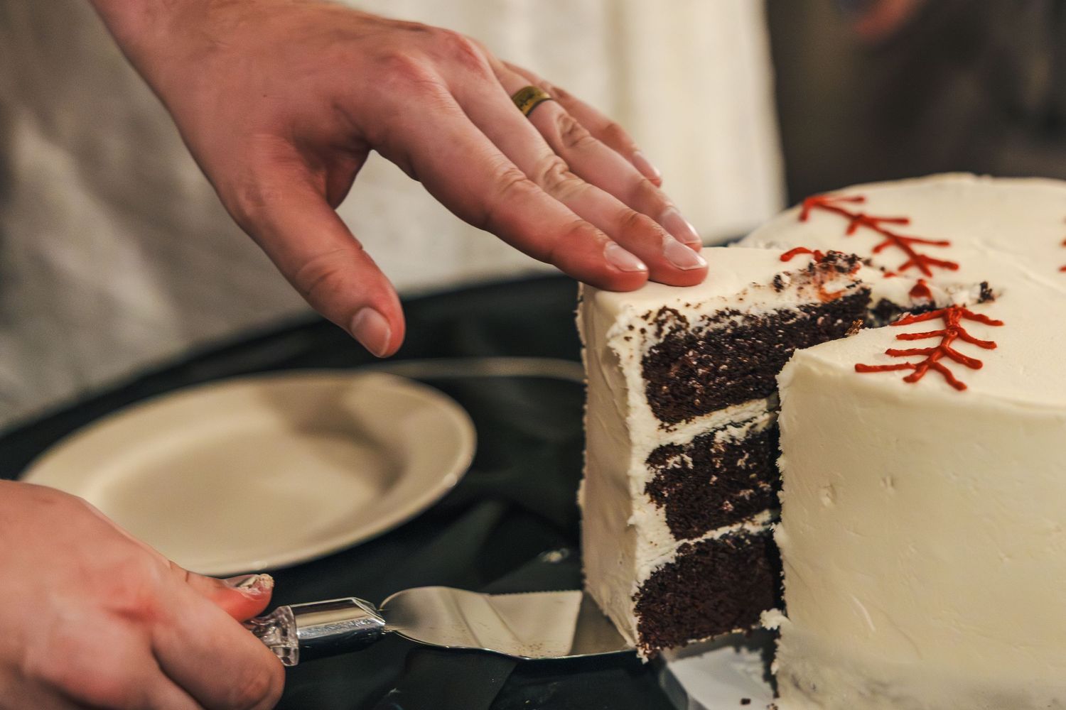 Hands slice into a white frosted chocolate cake decorated with red stitching to look like a baseball.