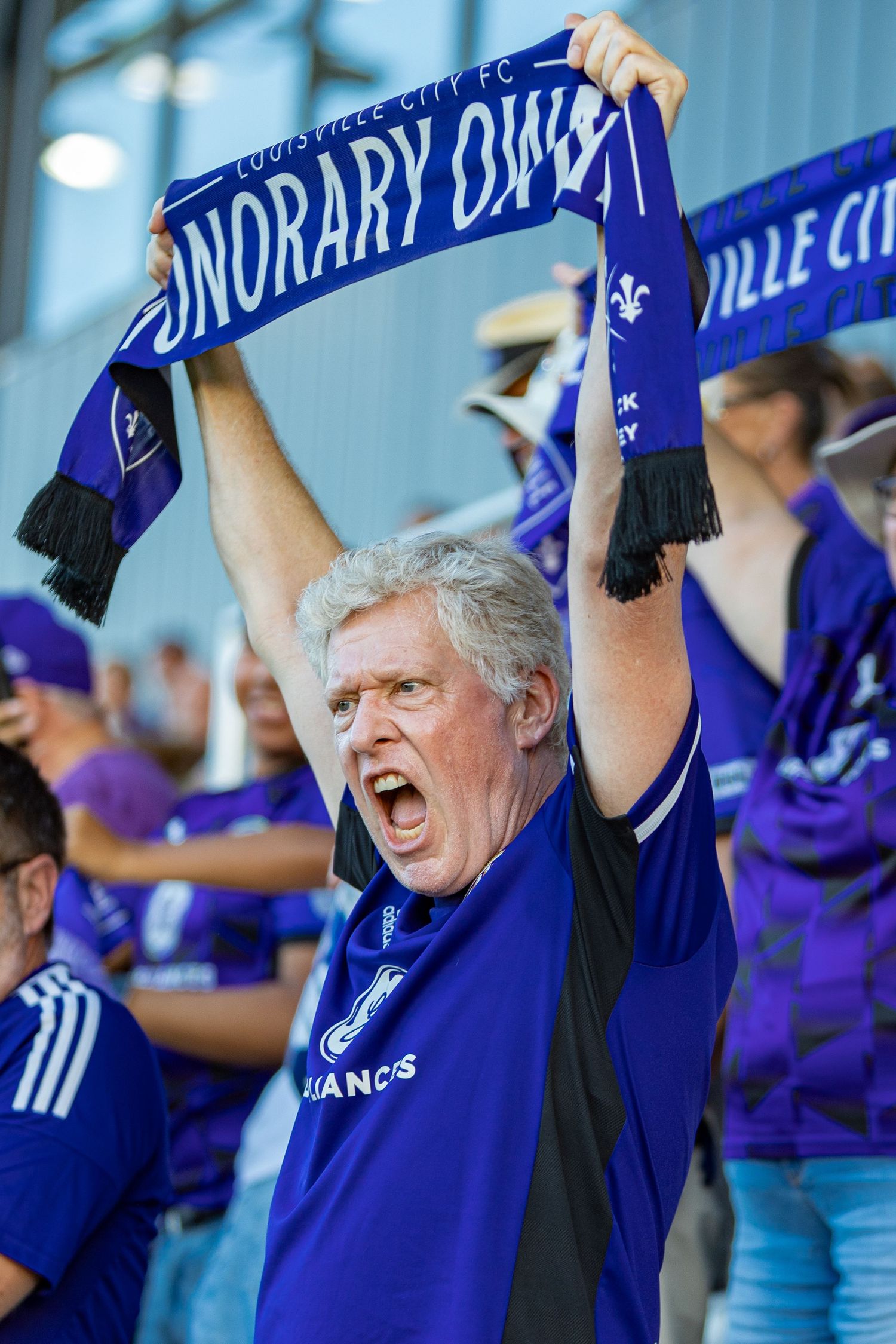 Passionate sports fan in blue jersey holds up team scarf while cheering enthusiastically at a stadium.