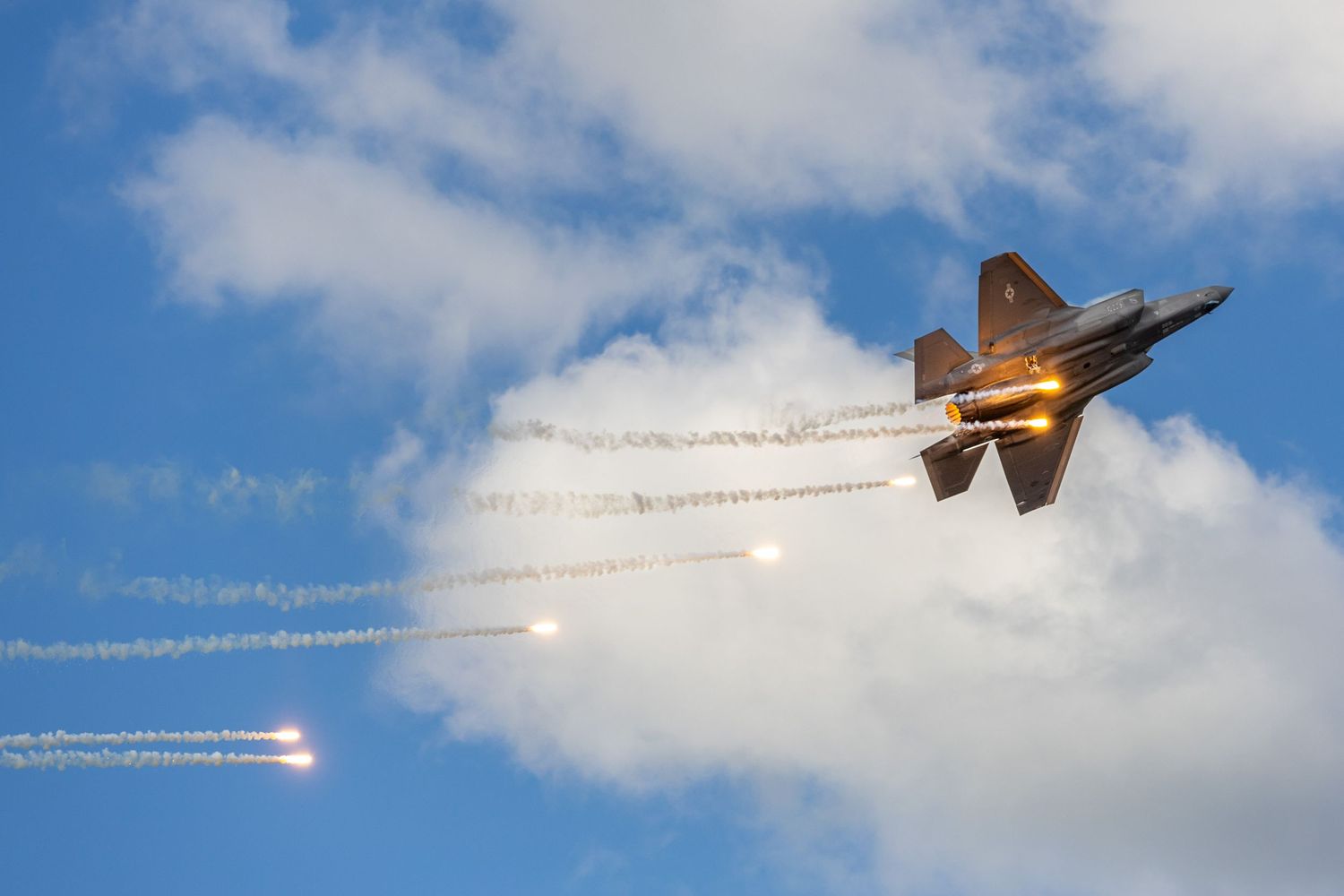 Fighter jet releasing flares while flying through cloudy sky during aerial demonstration.