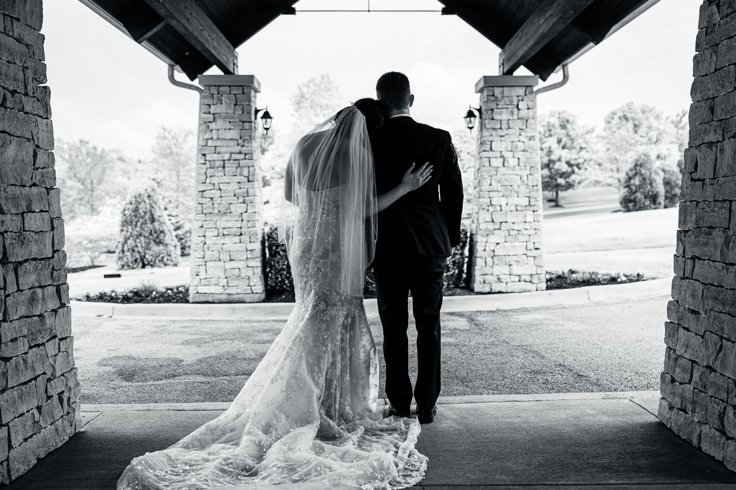 Black and white photo of bride and groom standing under stone archway, overlooking lush green golf course.