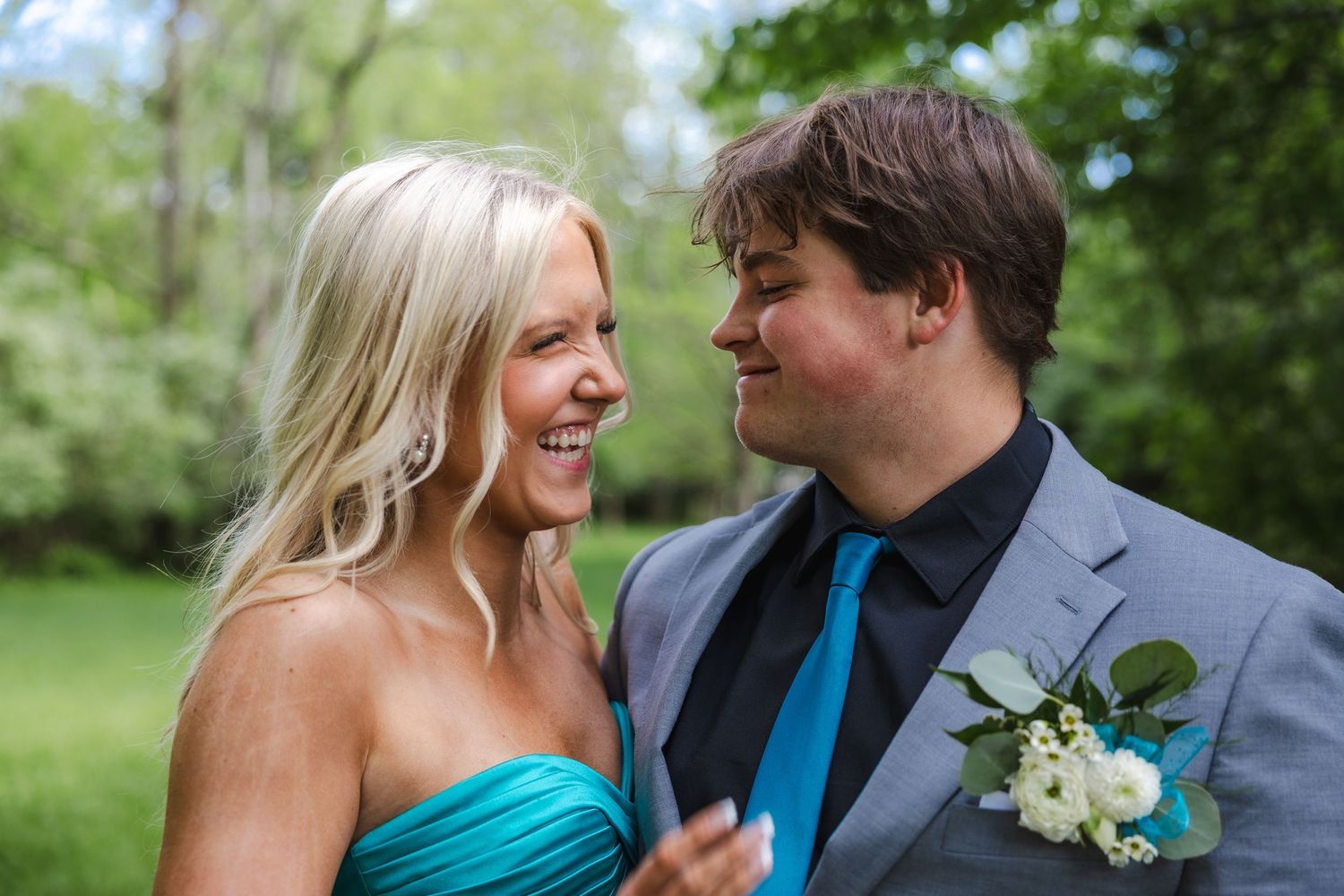 A couple shares a joyful moment outdoors, with her wearing a teal dress and him in a grey suit.