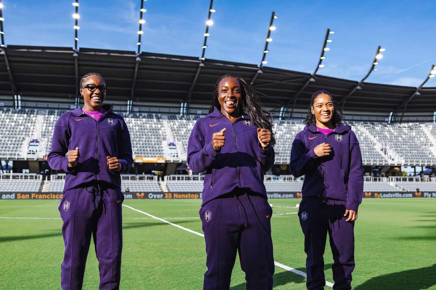 Three people in purple athletic wear pose together on a soccer field at a stadium.