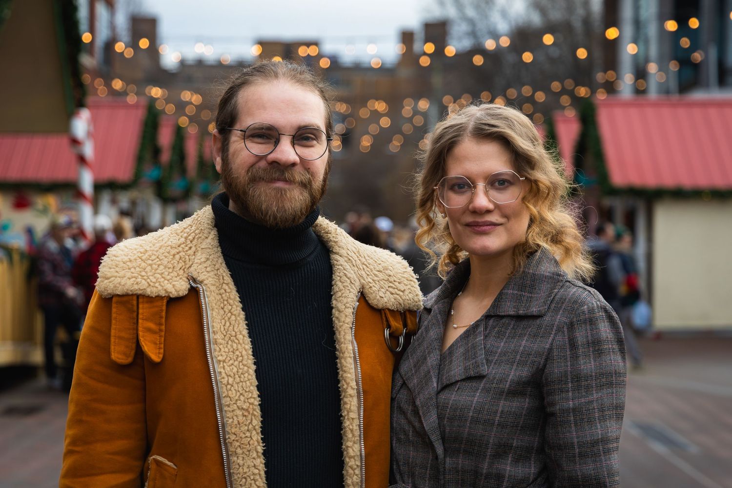 Couple in winter attire stands together at festive market with string lights behind.