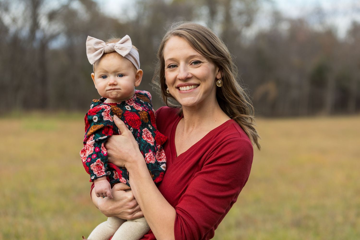 A woman in a burgundy sweater holds a baby wearing a floral outfit and bow headband in a field during autumn.