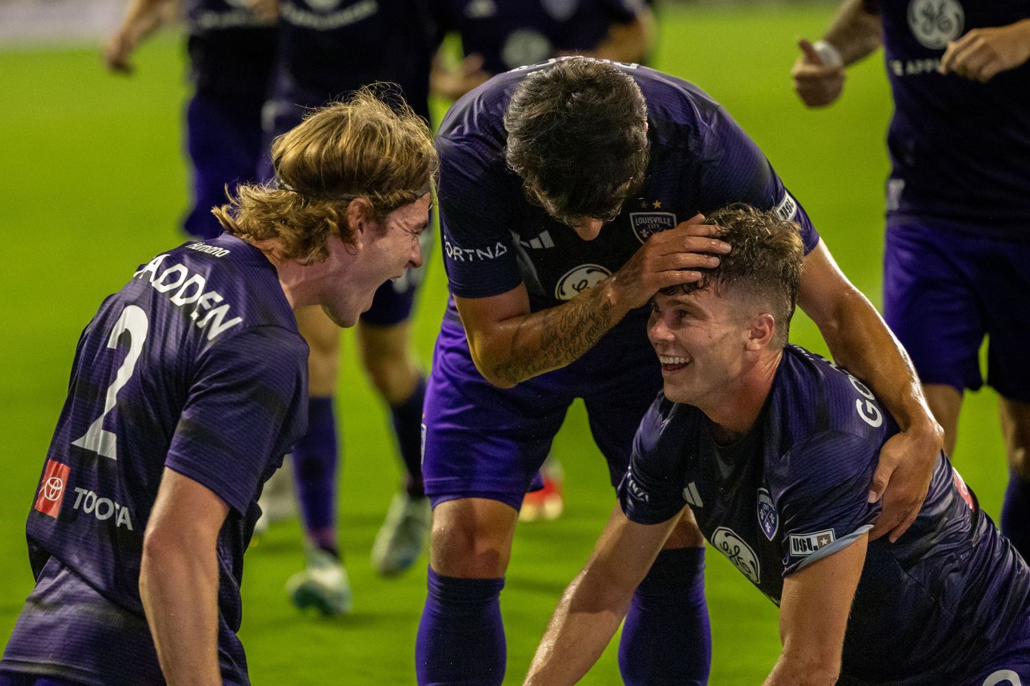 Team huddle during soccer match shows players celebrating together in purple uniforms.