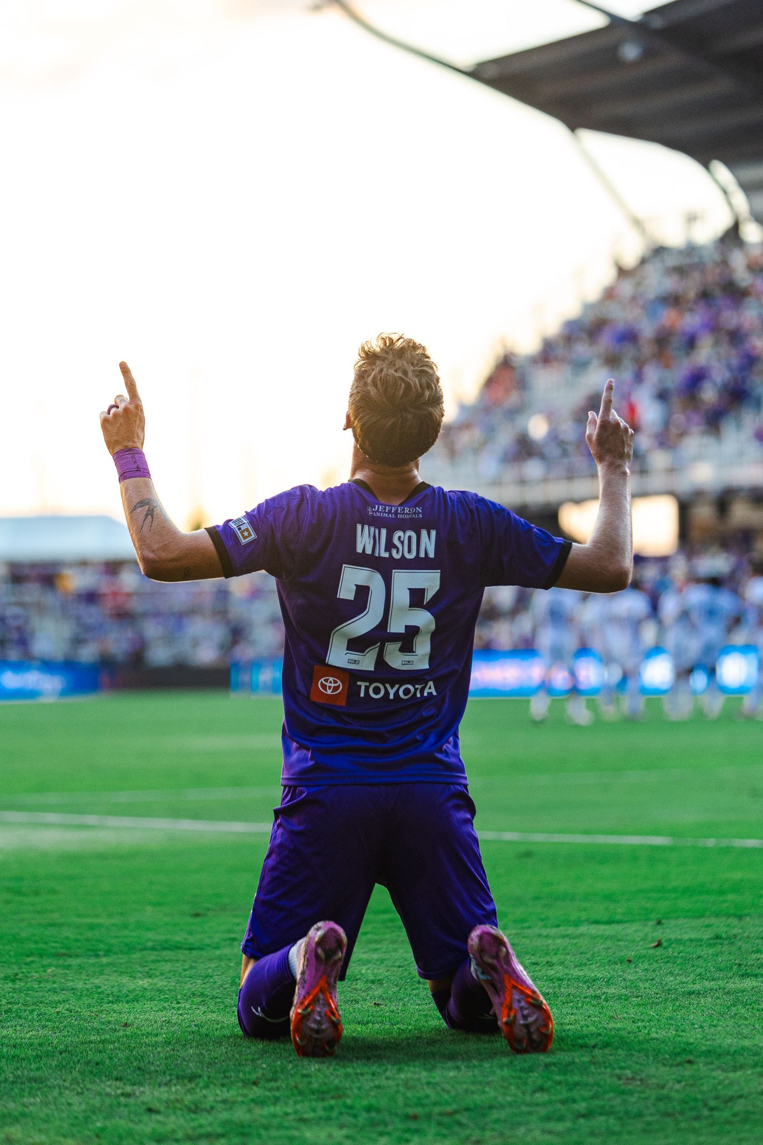 Soccer player in purple jersey kneels on grass field with arms raised in celebration during match.