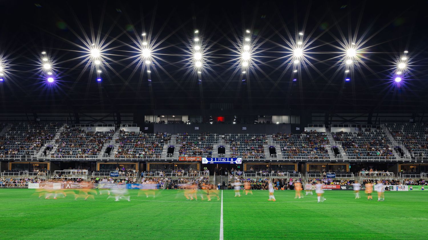 Panoramic view of a soccer stadium at night with bright floodlights illuminating the playing field and stands.