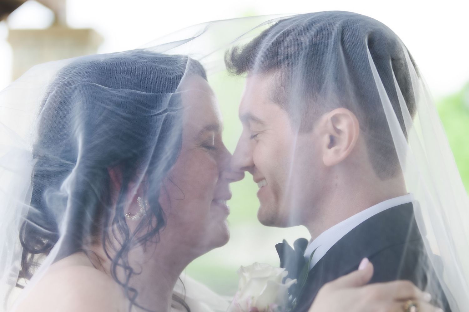 Bride and groom sharing an intimate moment under a flowing wedding veil, nearly kissing at their ceremony.