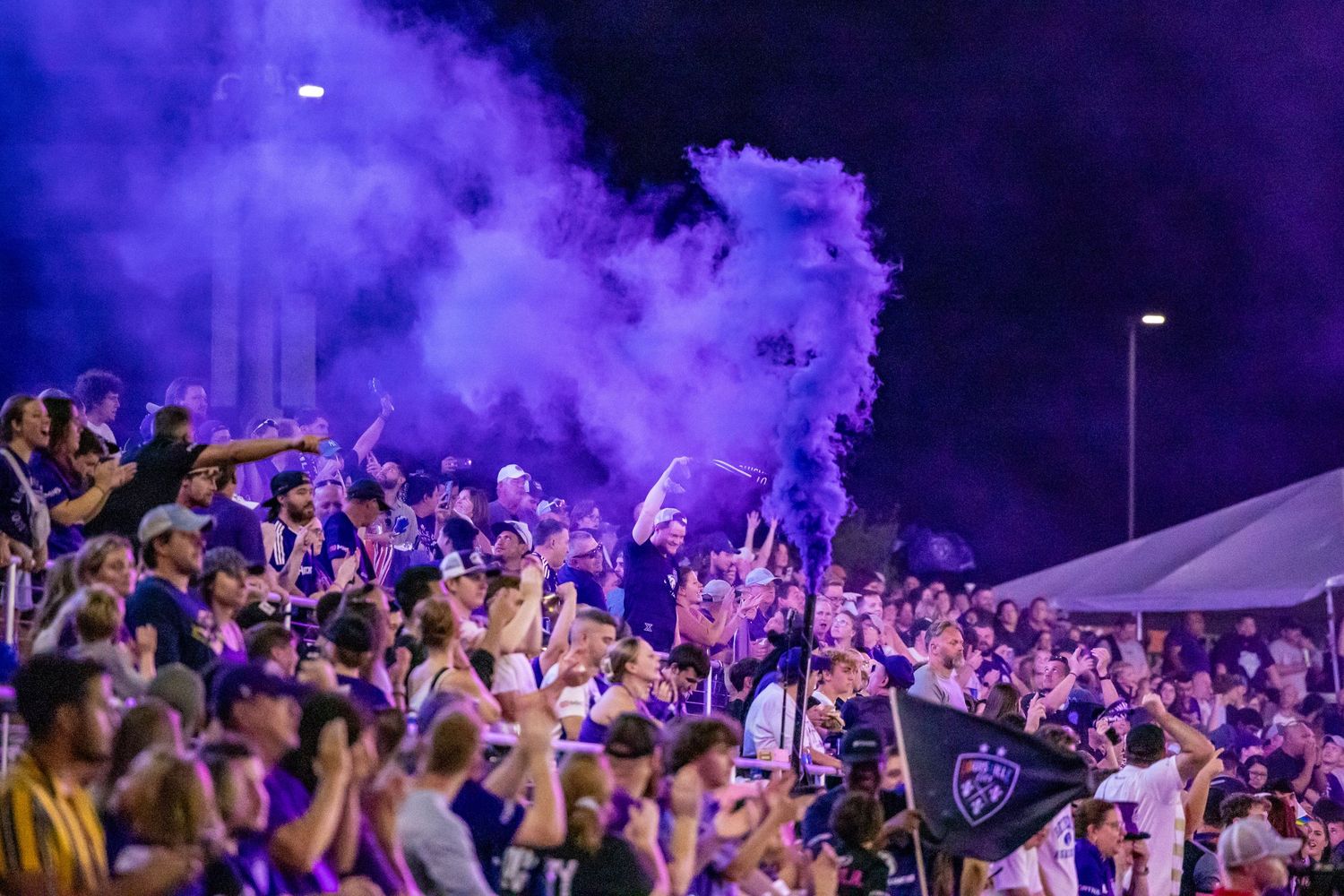 Enthusiastic soccer fans create a vibrant atmosphere with purple smoke and cheering in a crowded stadium section.