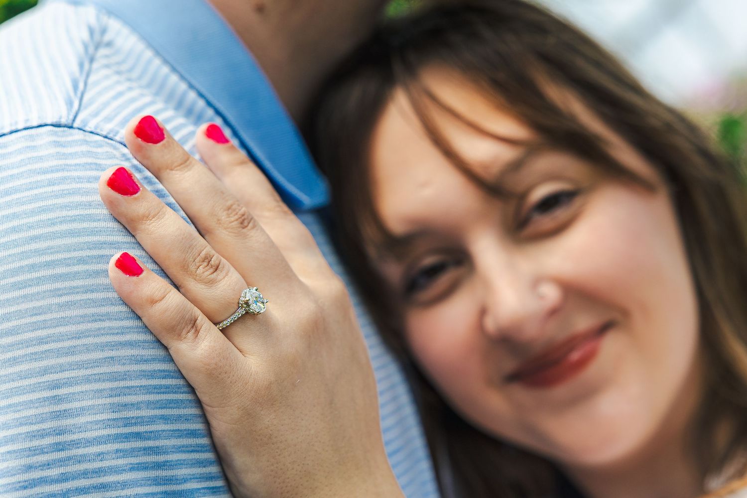Woman smiling with red nails and diamond engagement ring resting on man's shoulder in romantic outdoor photo.