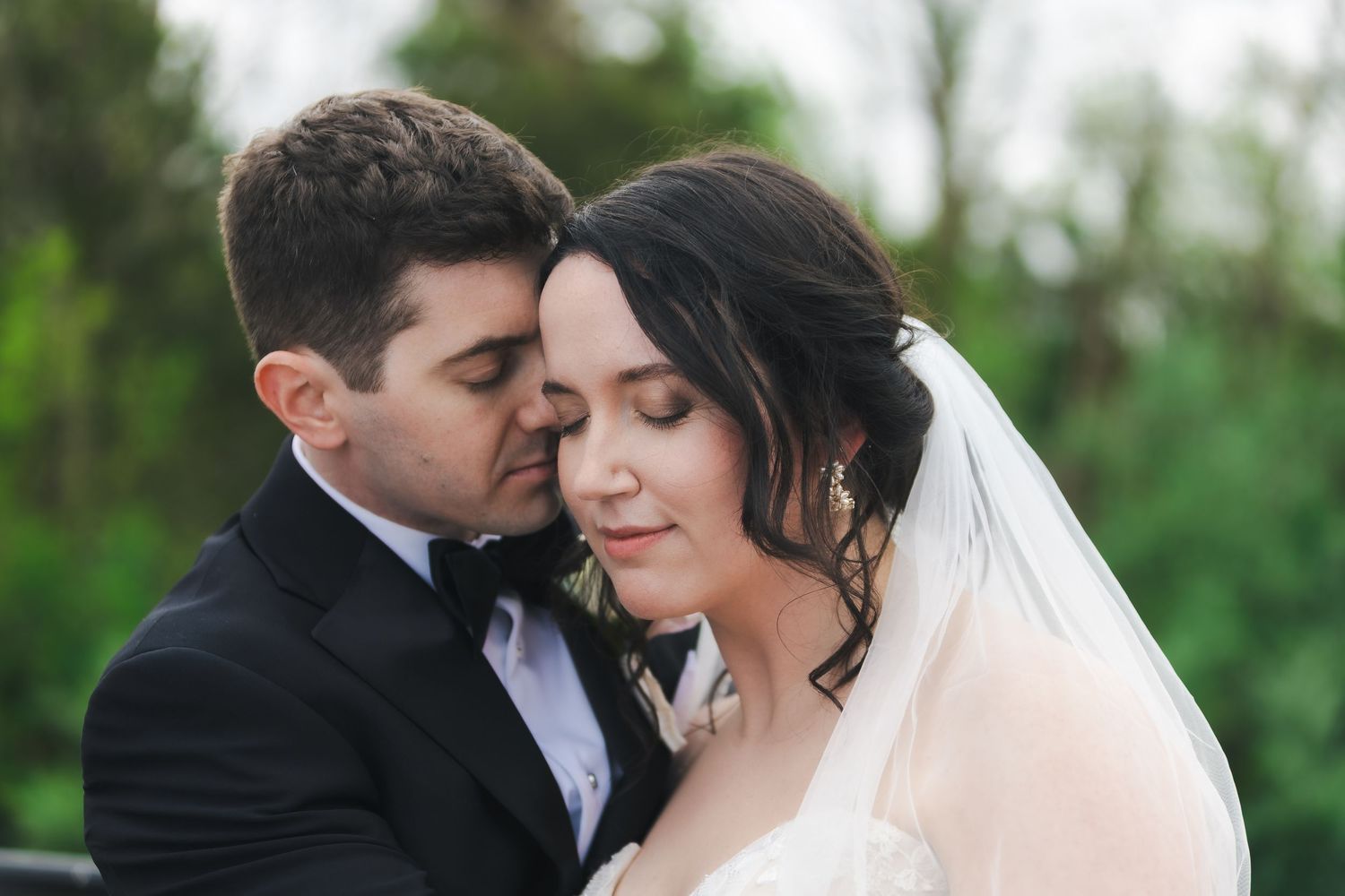 A groom in a black tuxedo tenderly nuzzles his bride in a white wedding gown and veil outdoors.