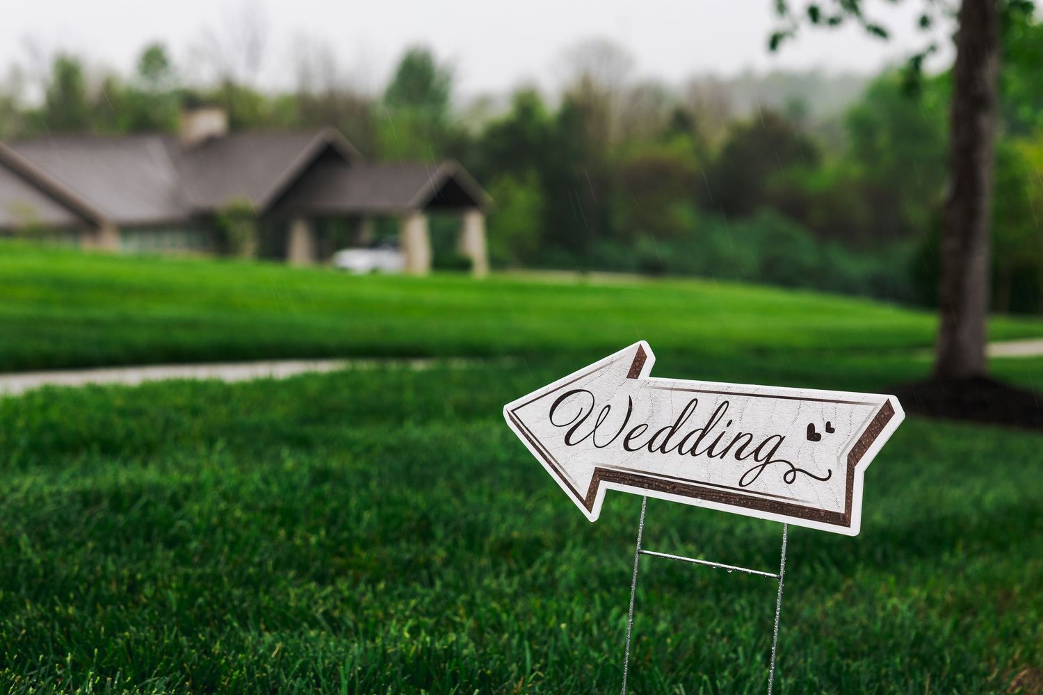 Arrow-shaped wedding directional sign on green grass lawn with a house and trees in the background.