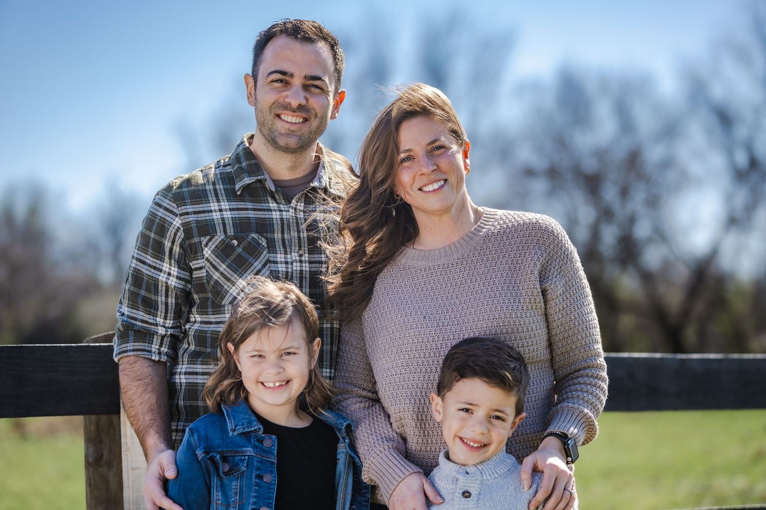 Happy family of four poses together on wooden bench during outdoor photo session.