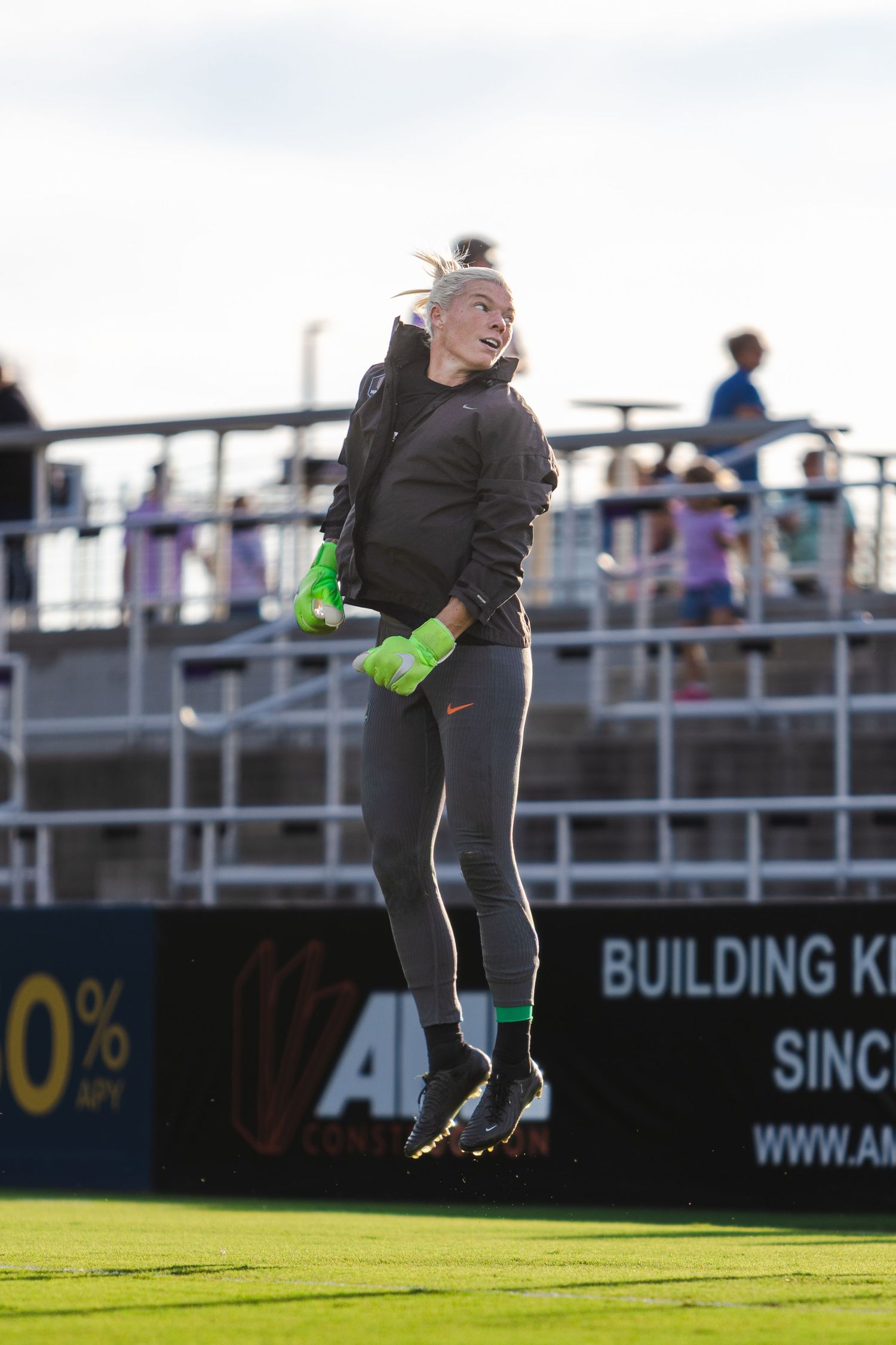 A soccer goalkeeper in black uniform and green gloves practices diving saves on a sunny field during training.