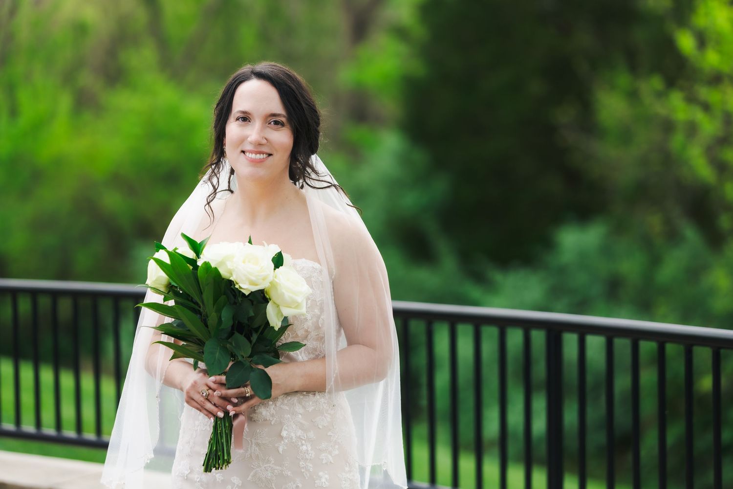 Smiling bride in lace gown holding white rose bouquet stands on outdoor deck surrounded by lush greenery.