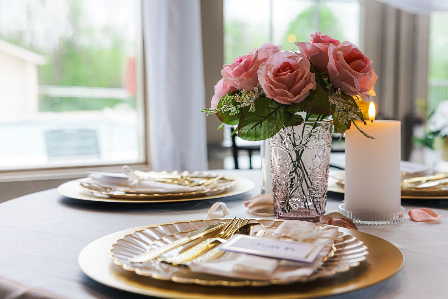 Elegant wedding table setting with pink roses, gold charger plates, and golden cutlery near a sunlit window.