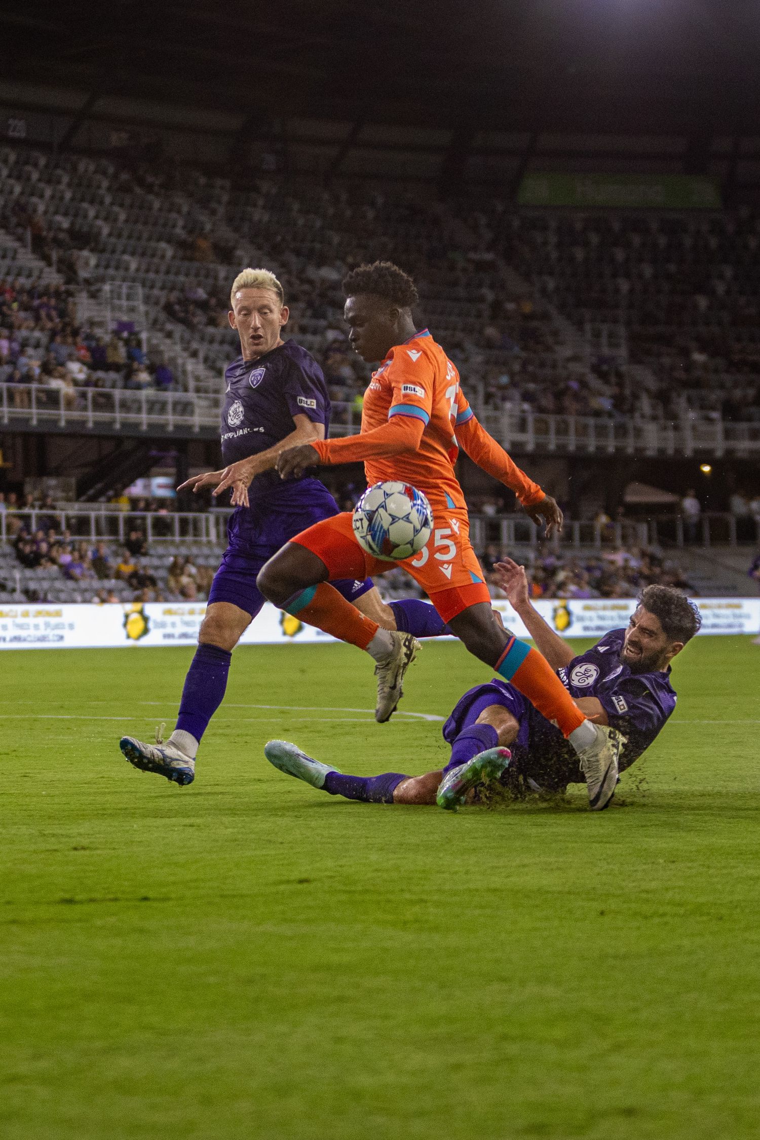 Soccer players from opposing teams battle for possession during competitive match on illuminated field.