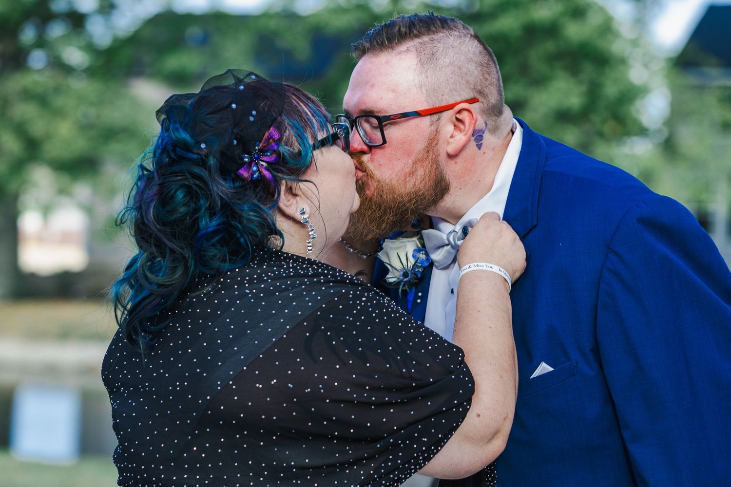 A couple shares a romantic kiss during their wedding ceremony against a lush green outdoor backdrop.