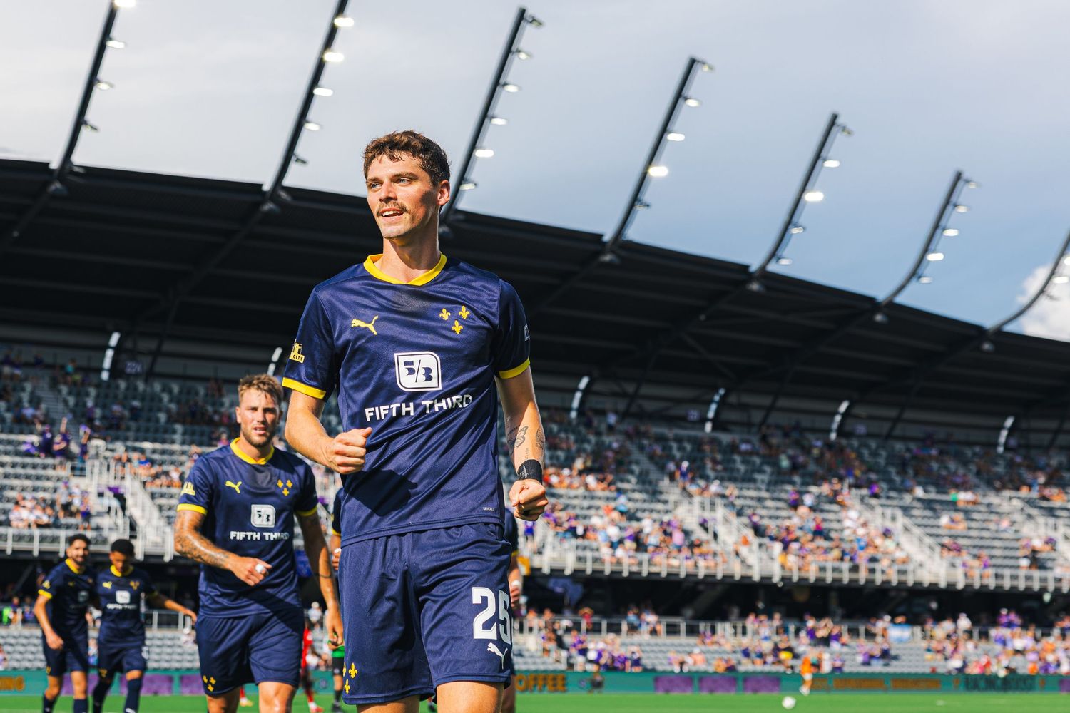 Soccer player in navy blue jersey celebrates during match at stadium with empty seats in background.