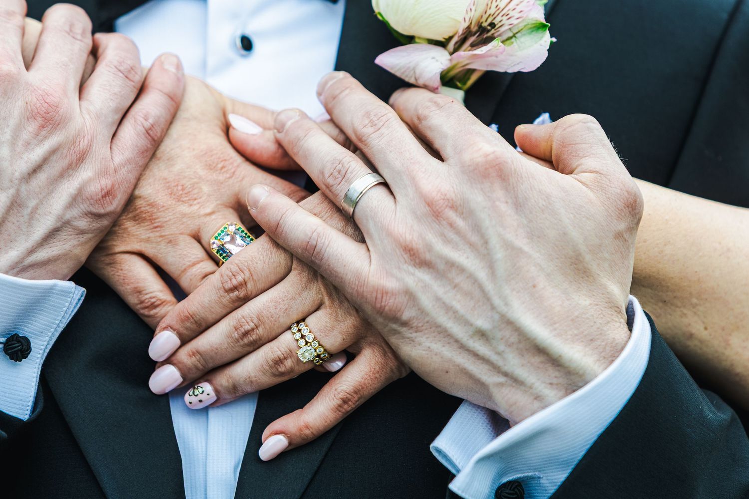 Close-up of bride and groom hands showing wedding rings on black tuxedo jacket.