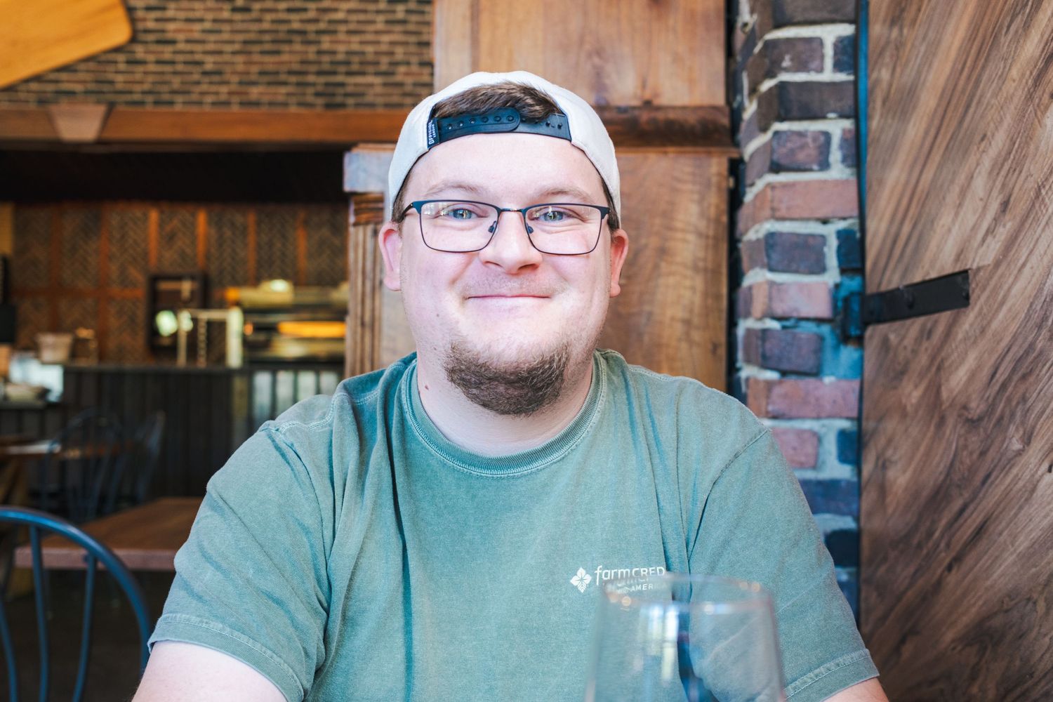 Smiling young man wearing glasses and a backwards cap sitting in a rustic brick restaurant.