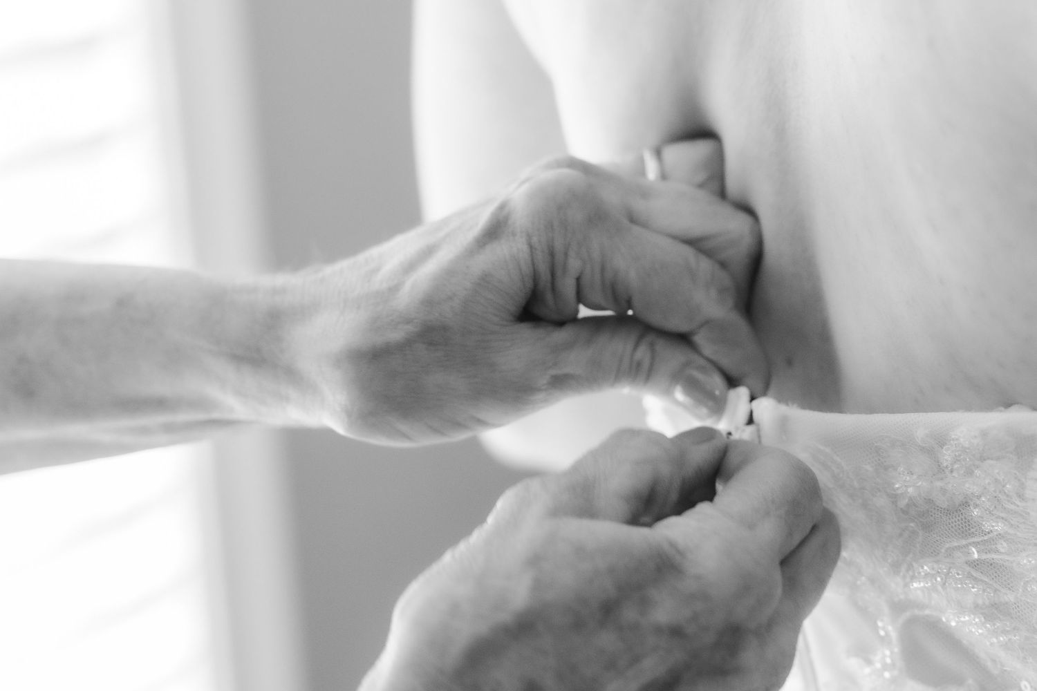 Close-up black and white photo of hands fastening buttons on a wedding dress during bridal preparations.