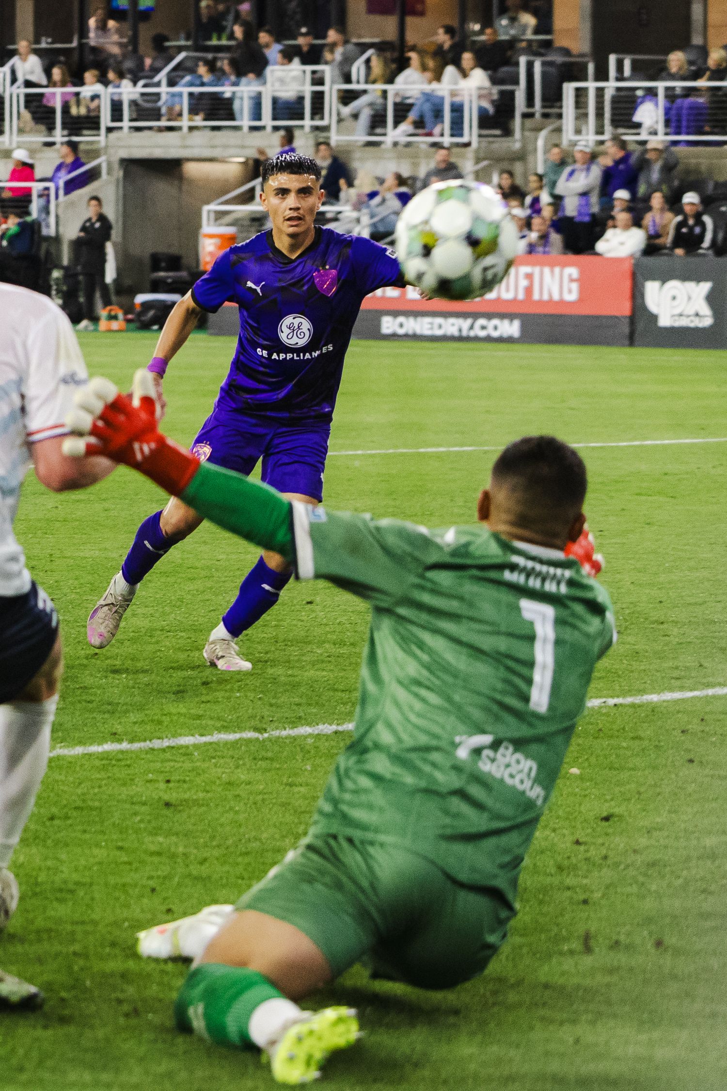 Goalkeeper slides to make a save as attacker in purple jersey approaches the ball.
