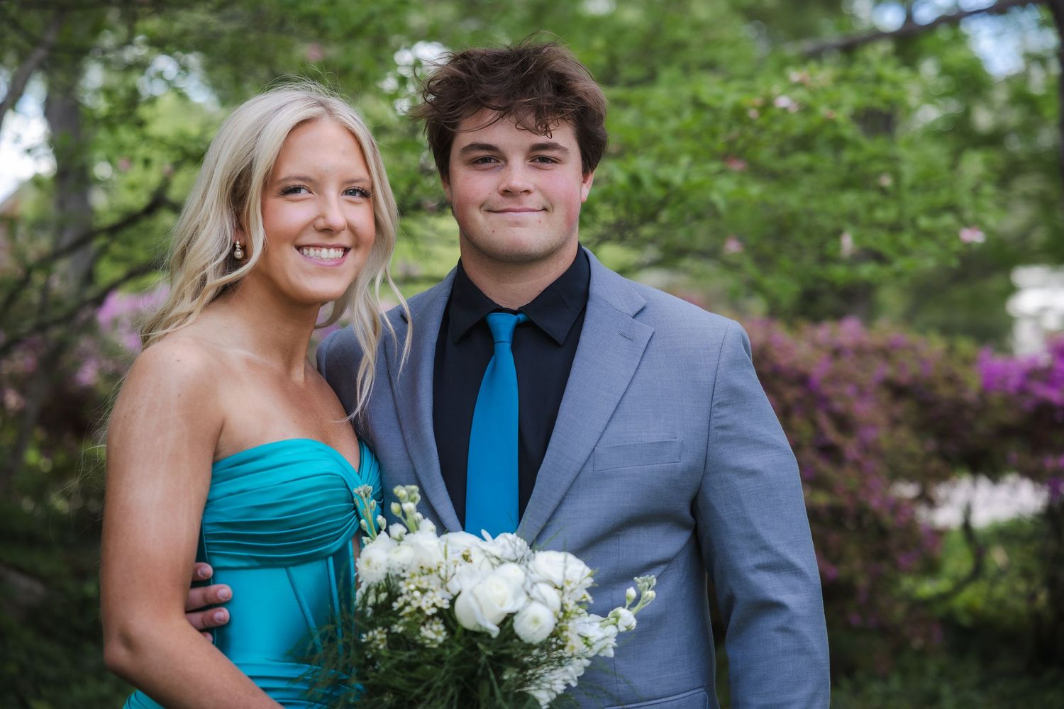 A happy couple in formal attire stands together outdoors with a bouquet of white flowers amid blooming spring foliage.